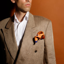 A man dressed in a beige suit, styled with the "Artist's Rhombs & Corals" unisex pochette square by FGTONSILK, a 100% Greek silk accessory in vibrant orange tones, set against a rich terracotta background.