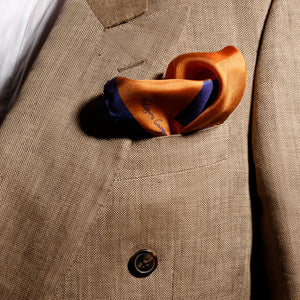 A man dressed in a beige suit, styled with the "Artist's Rhombs & Corals" unisex pochette square by FGTONSILK, a 100% Greek silk accessory in vibrant orange tones, set against a rich terracotta background.